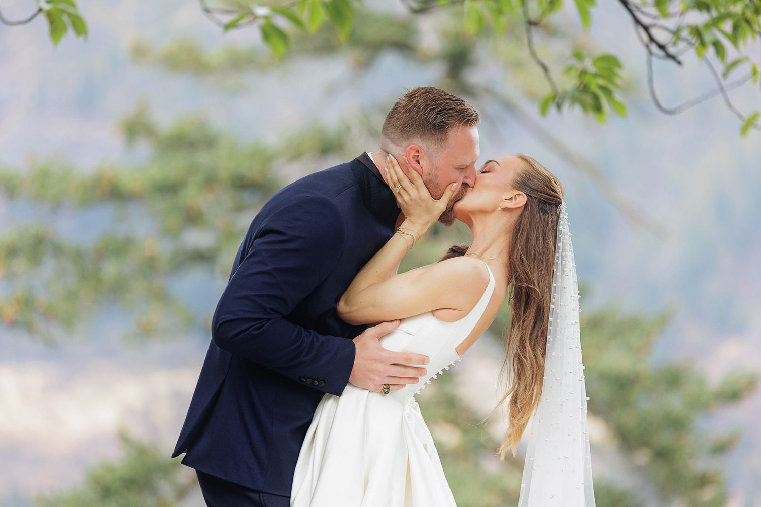 Bride and groom share a romantic kiss outdoors, bride in white gown with veil, groom in navy suit.