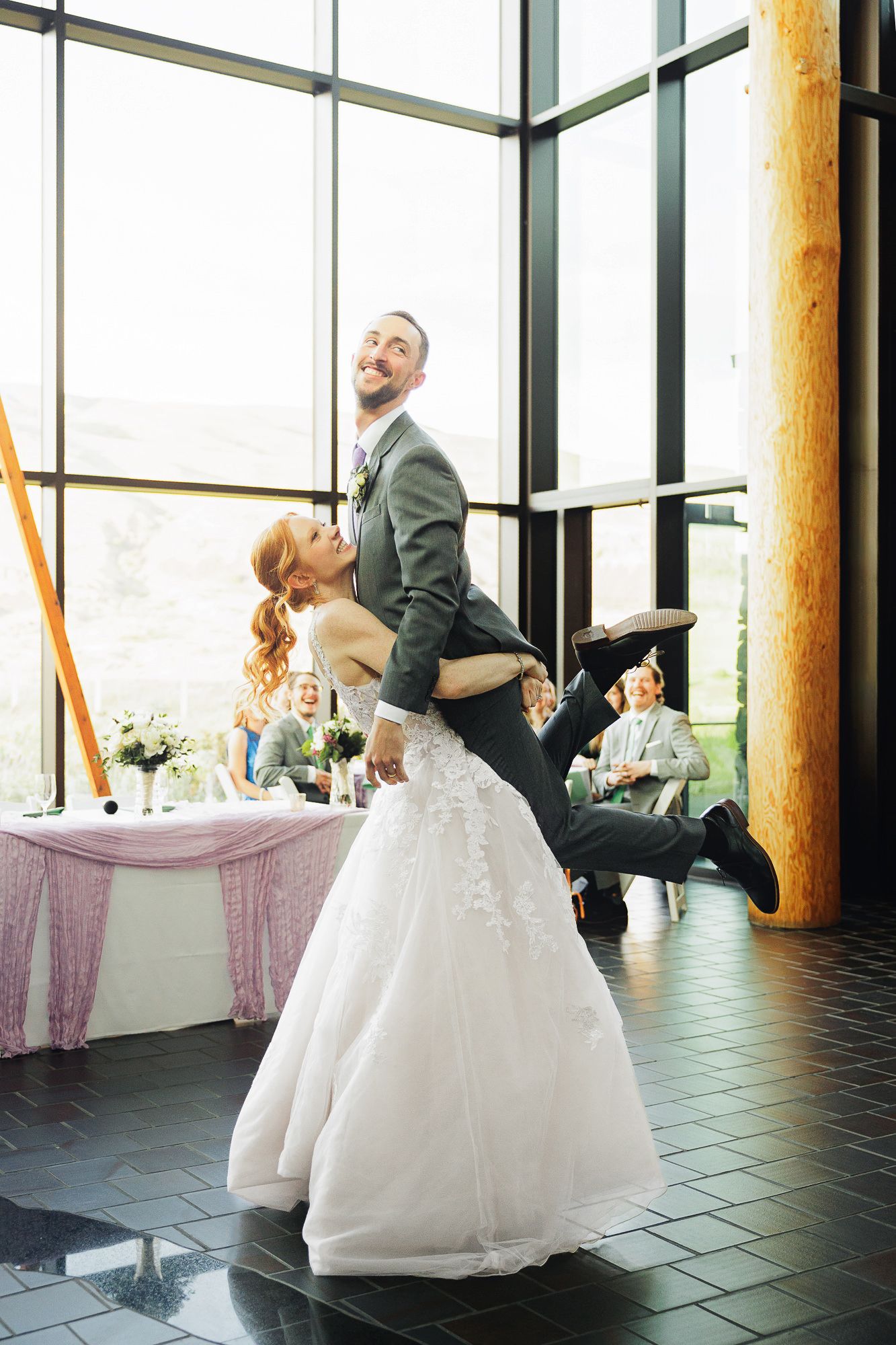 Bride lifts groom during first dance at elegant wedding reception with large windows and guests seated nearby.