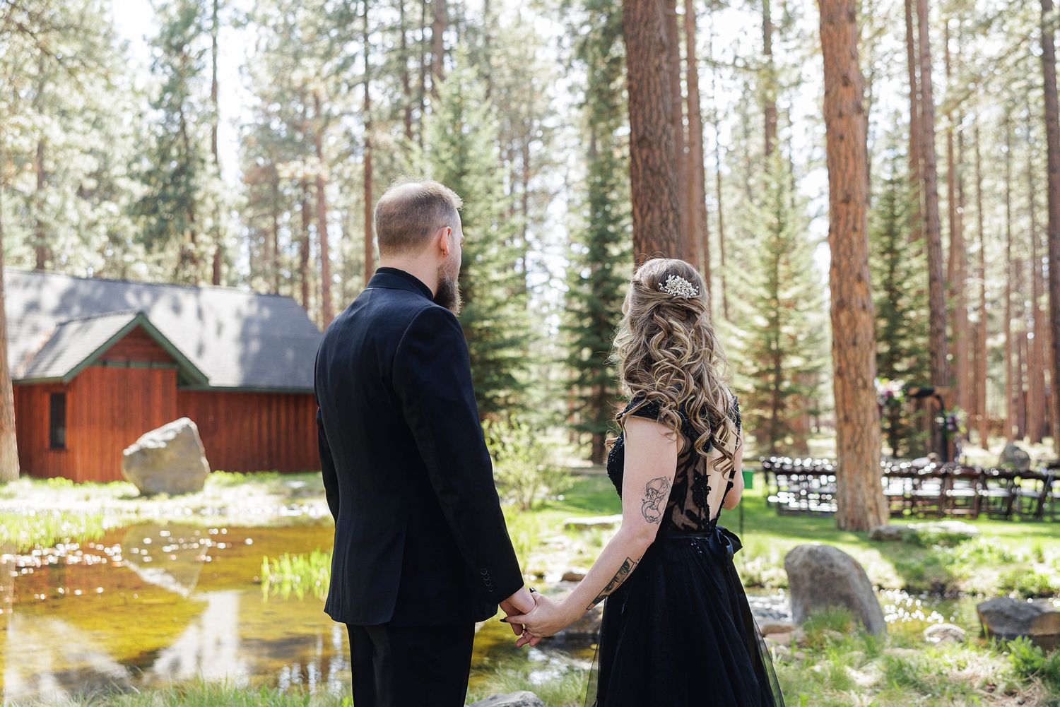 Couple holding hands in black formal wear among tall pine trees near a rustic red cabin in a forest setting.