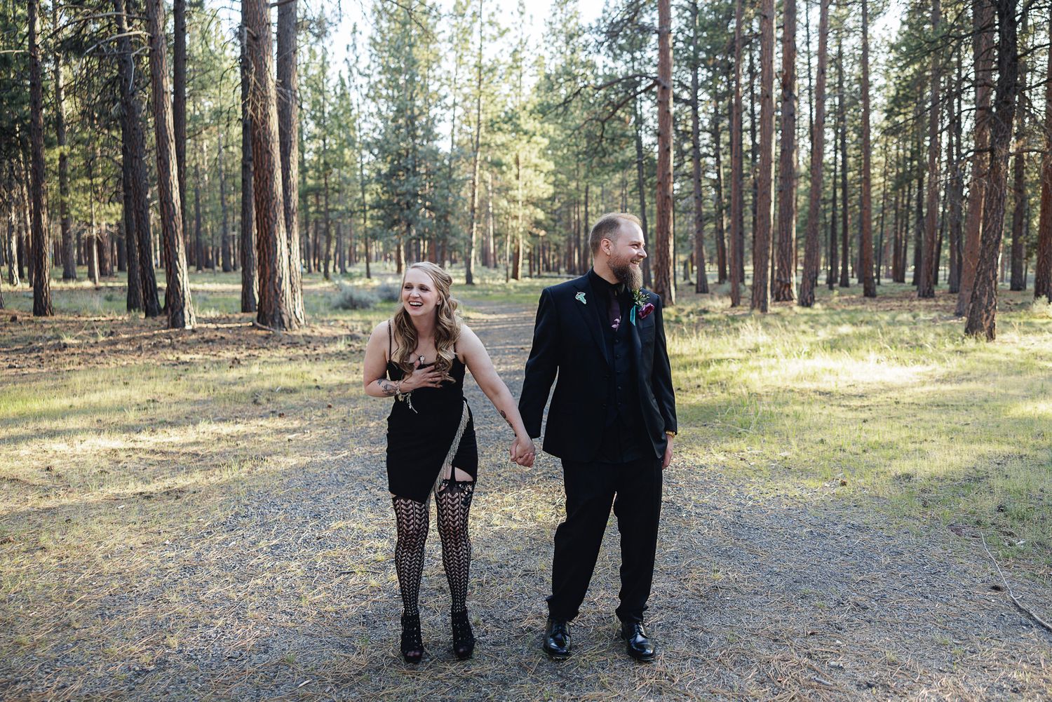 A couple holds hands walking through a pine forest, woman in black dress with fishnet stockings, man in black suit.