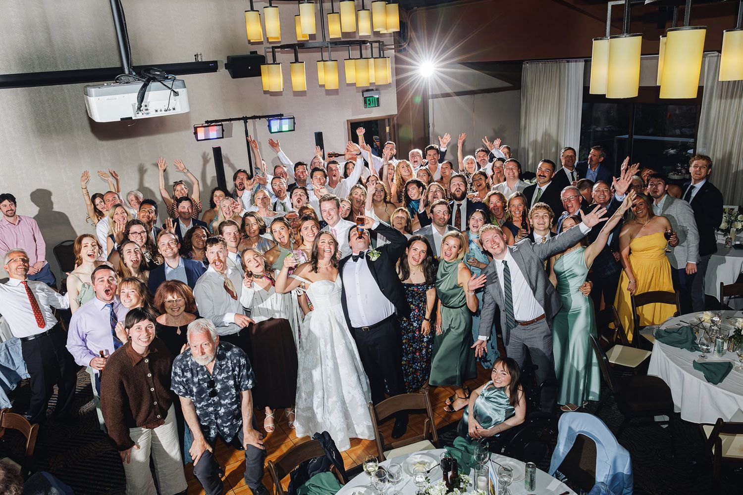 Large wedding party group photo with bride, groom, and guests cheering in decorated reception hall.