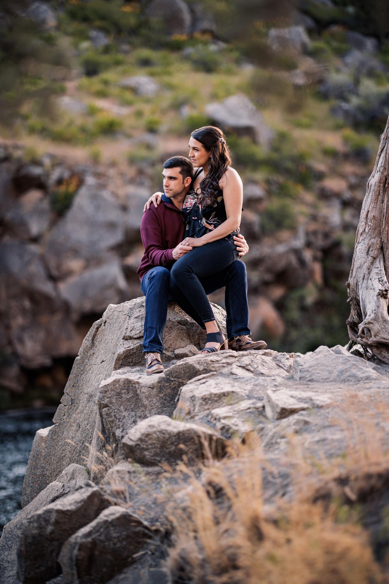 A couple sits together on rocky cliffs overlooking water during an outdoor photo session.