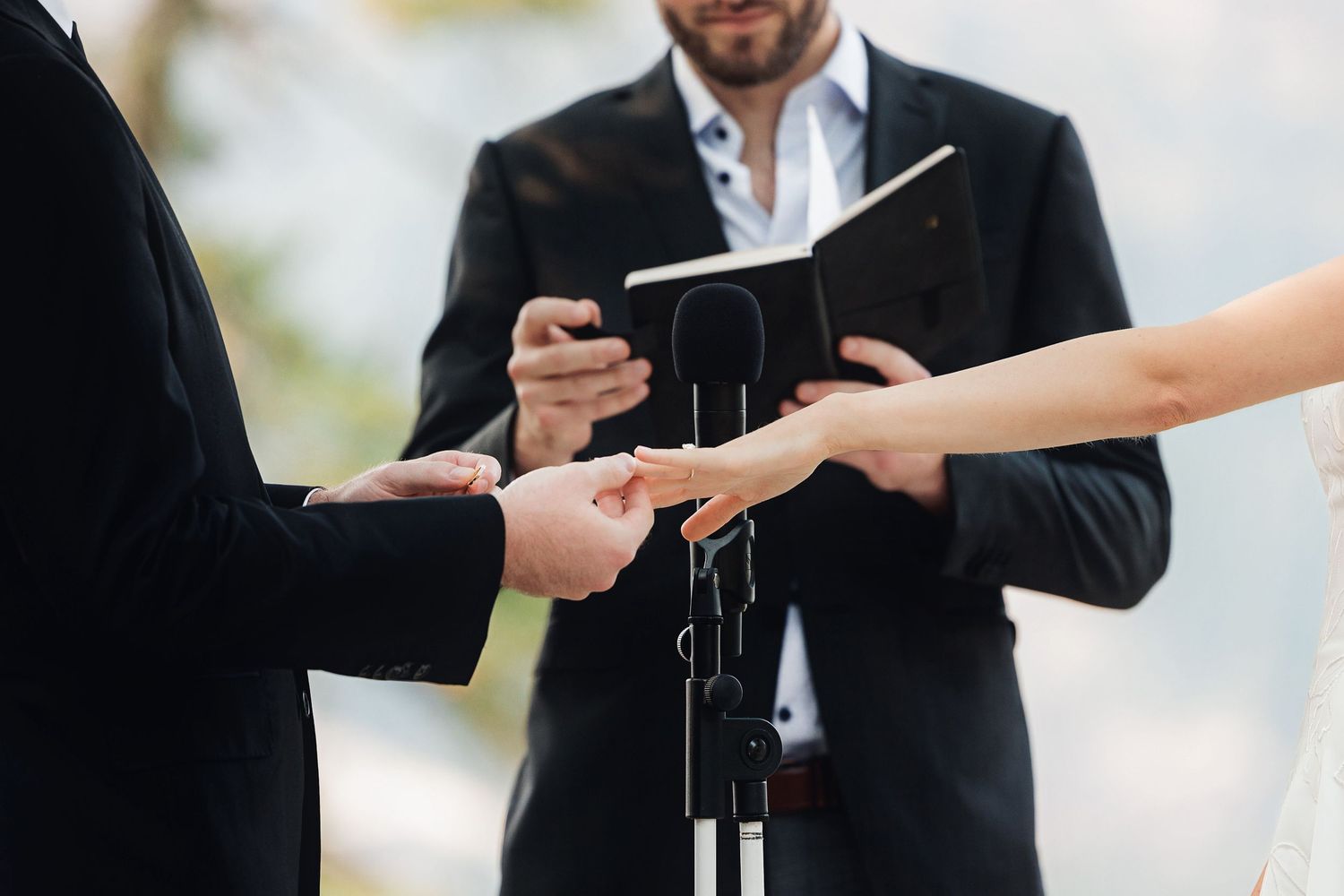 Couple exchanging rings during an outdoor wedding ceremony with an officiant holding a book.