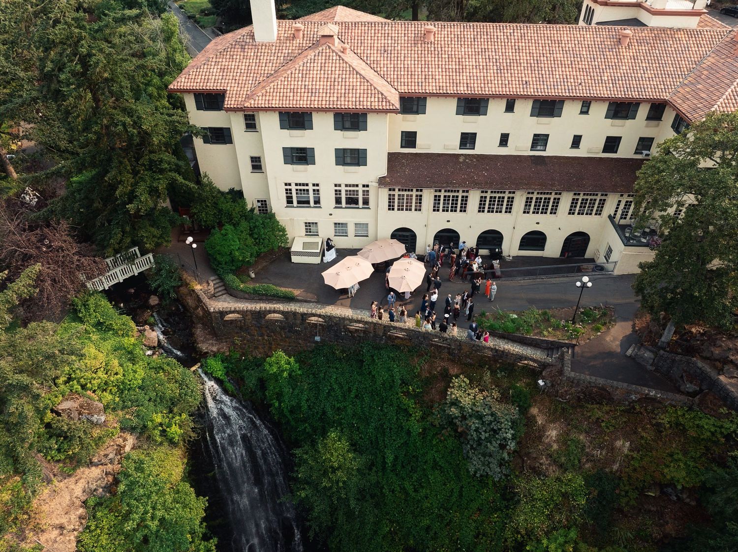 Aerial view of a historic hotel with terracotta roof near a waterfall, guests gathered under pink umbrellas on the patio.