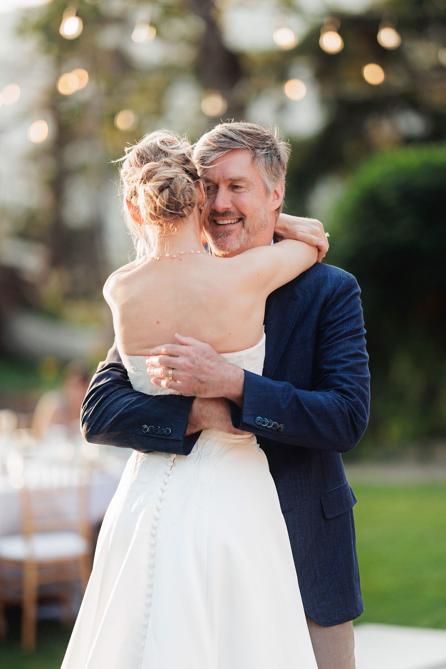 Father and bride share a joyful hug during an outdoor wedding reception with string lights in the background.