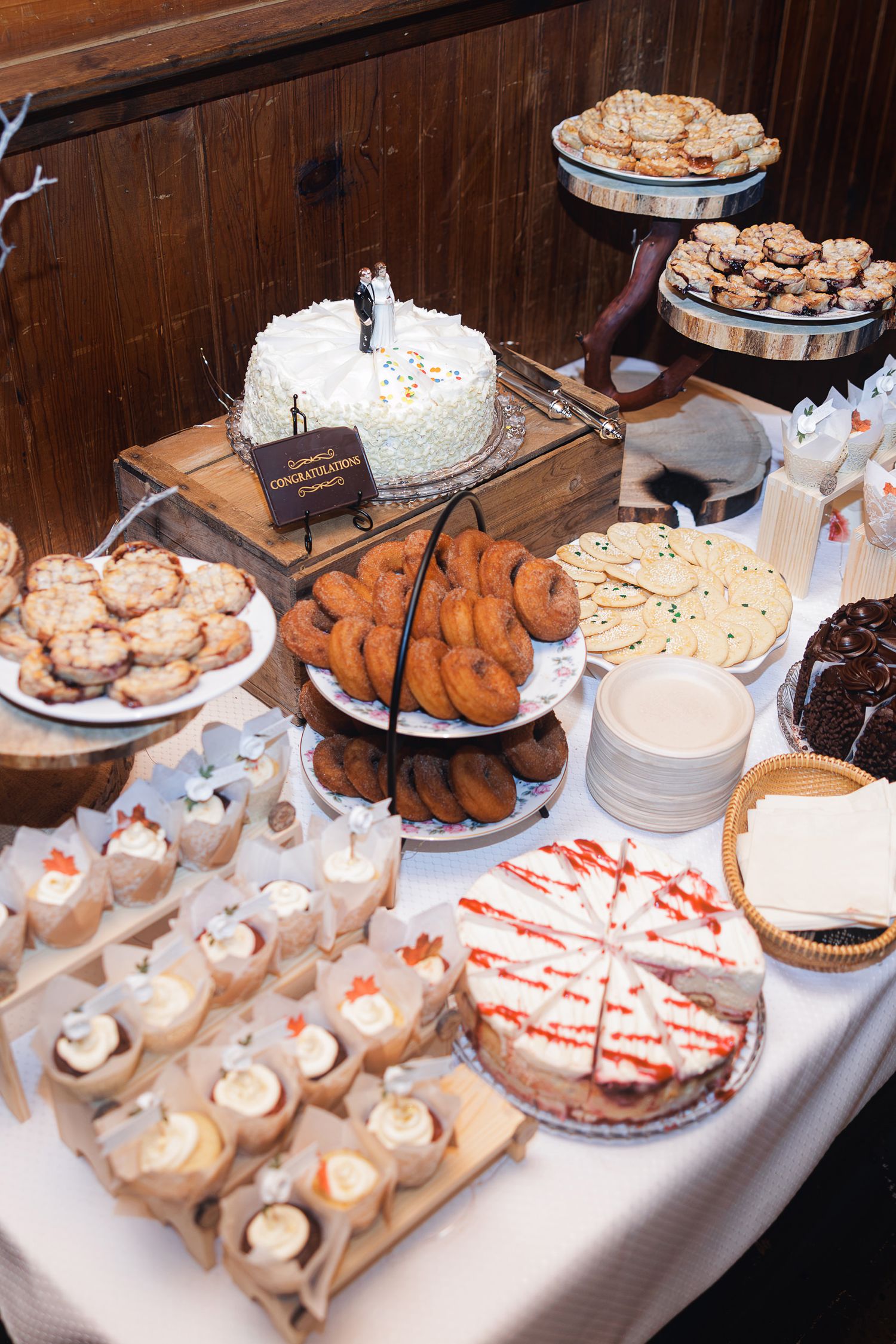 Elegant wedding dessert table with white cake, donuts, cupcakes, cookies, and cheesecake displayed on a white tablecloth.