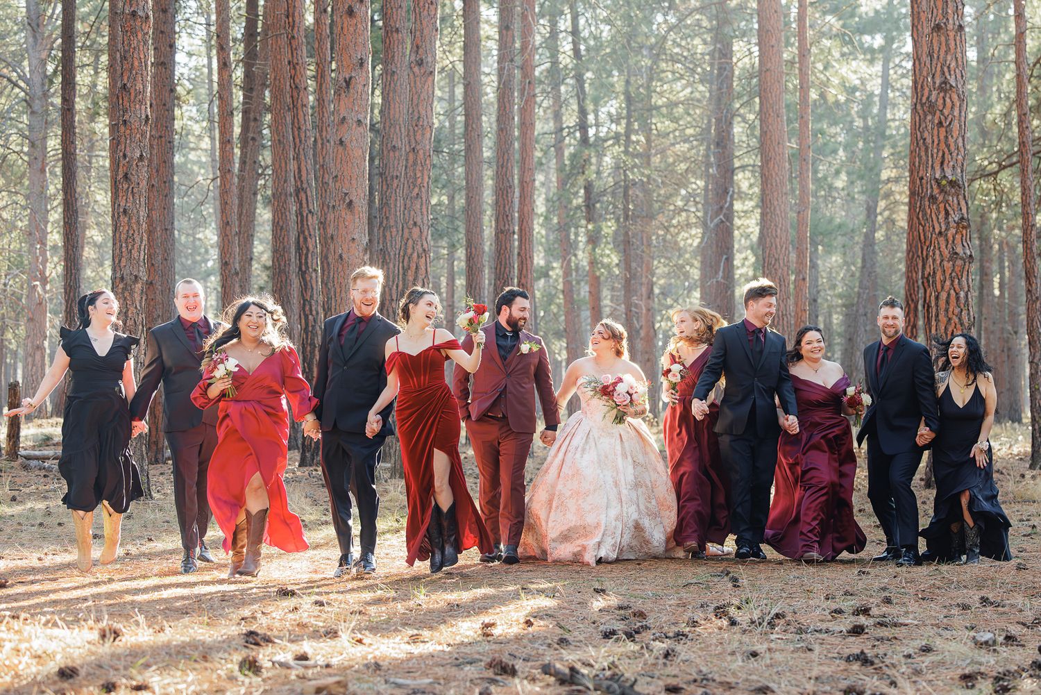 A large wedding party in burgundy and black attire poses together in a scenic forest setting with tall pine trees.