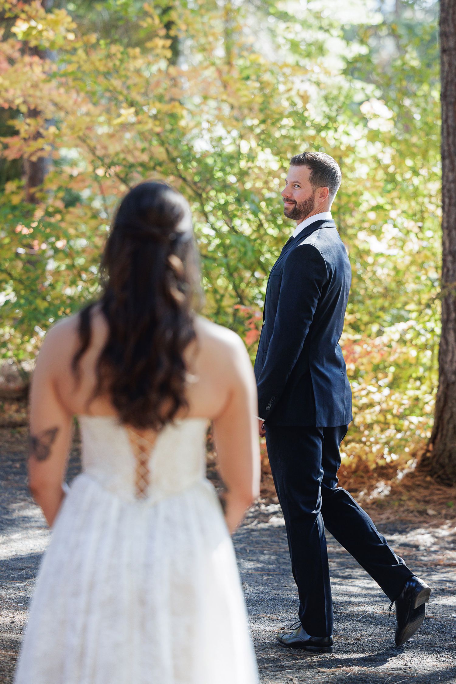 Groom in navy suit smiles at bride in lace wedding dress during outdoor first look among autumn trees.