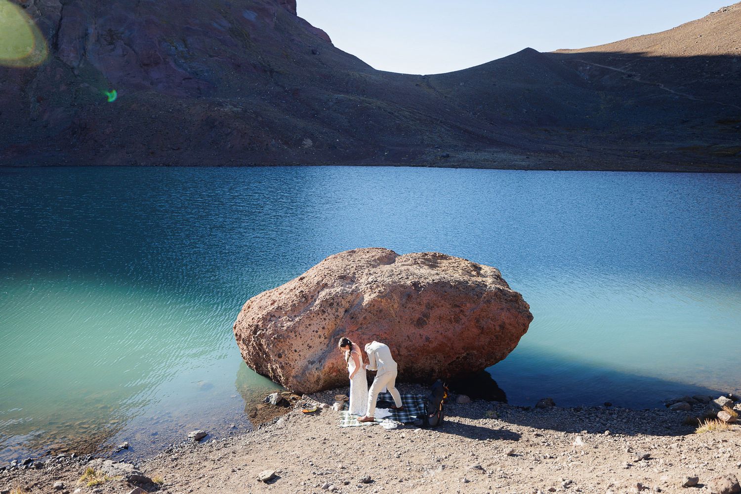 Hiker in white standing beside a large boulder on the rocky shore of a turquoise mountain lake with steep barren slopes behind.