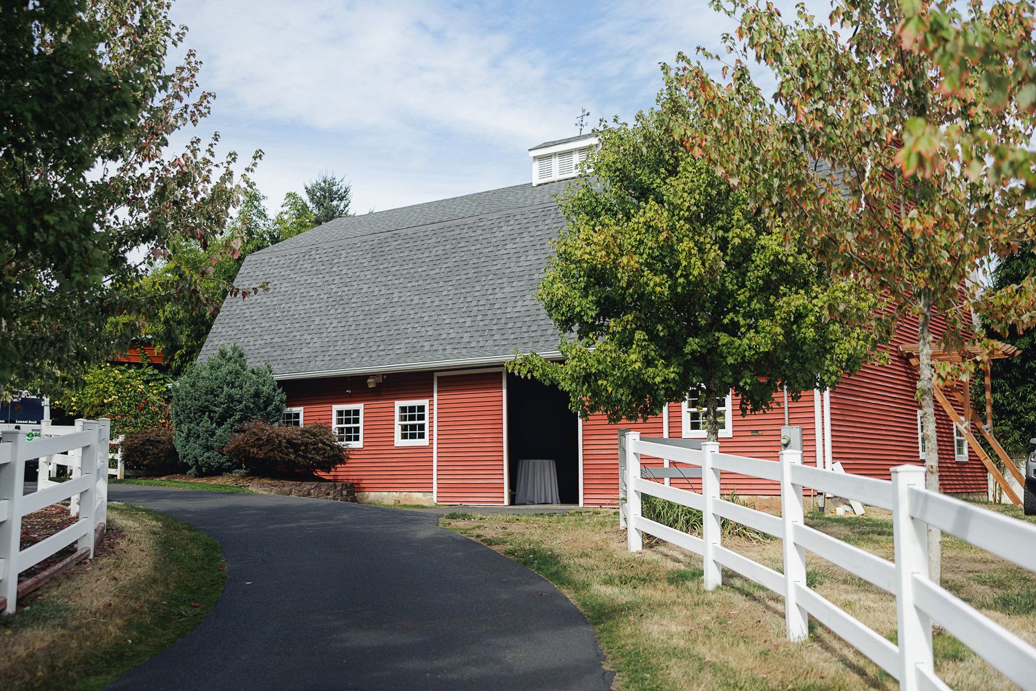 A panoramic view of a red barn with white fencing along a rural driveway on a sunny autumn day.