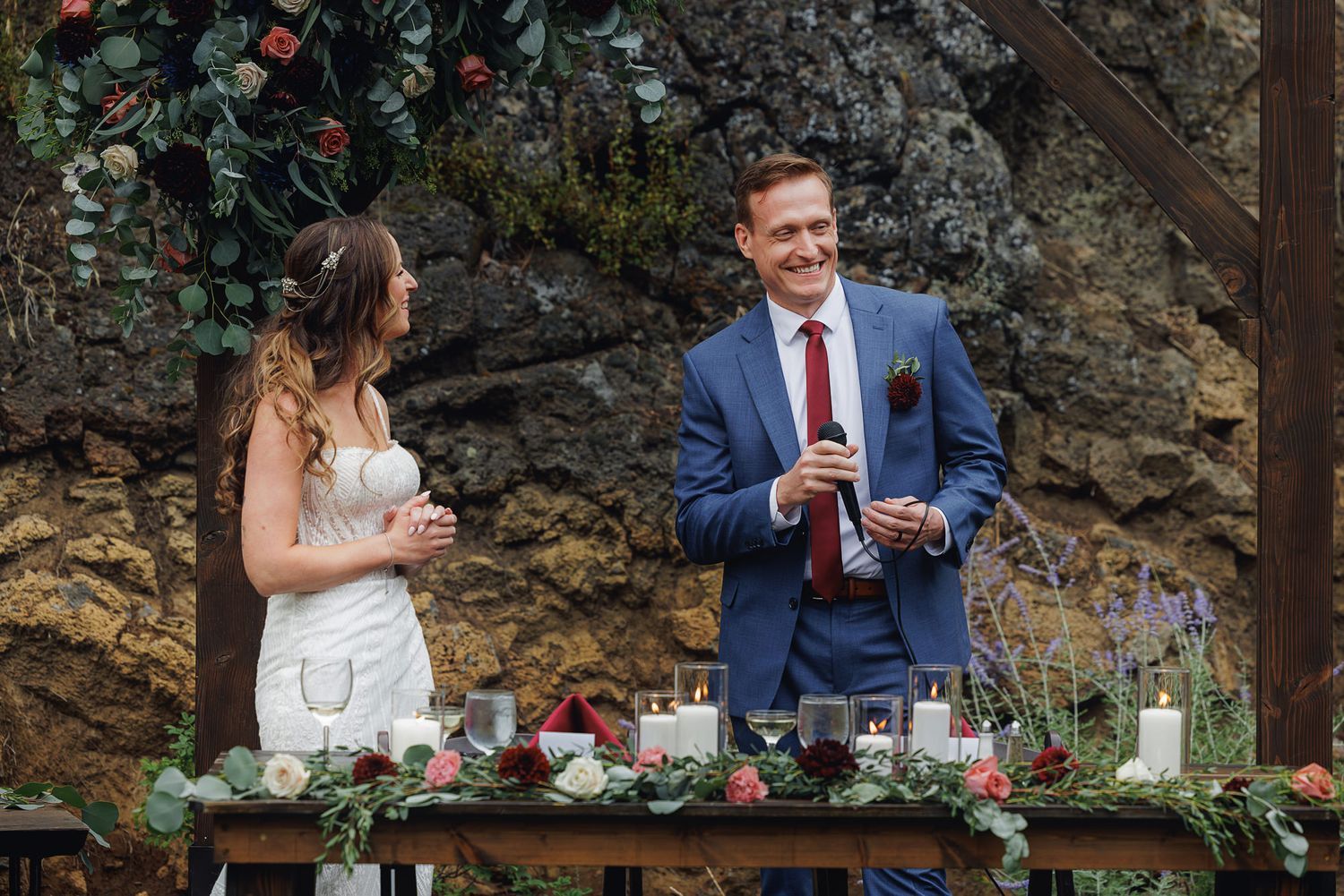 A couple shares a moment at their outdoor wedding reception with floral decorations and candles on a rustic wooden table.