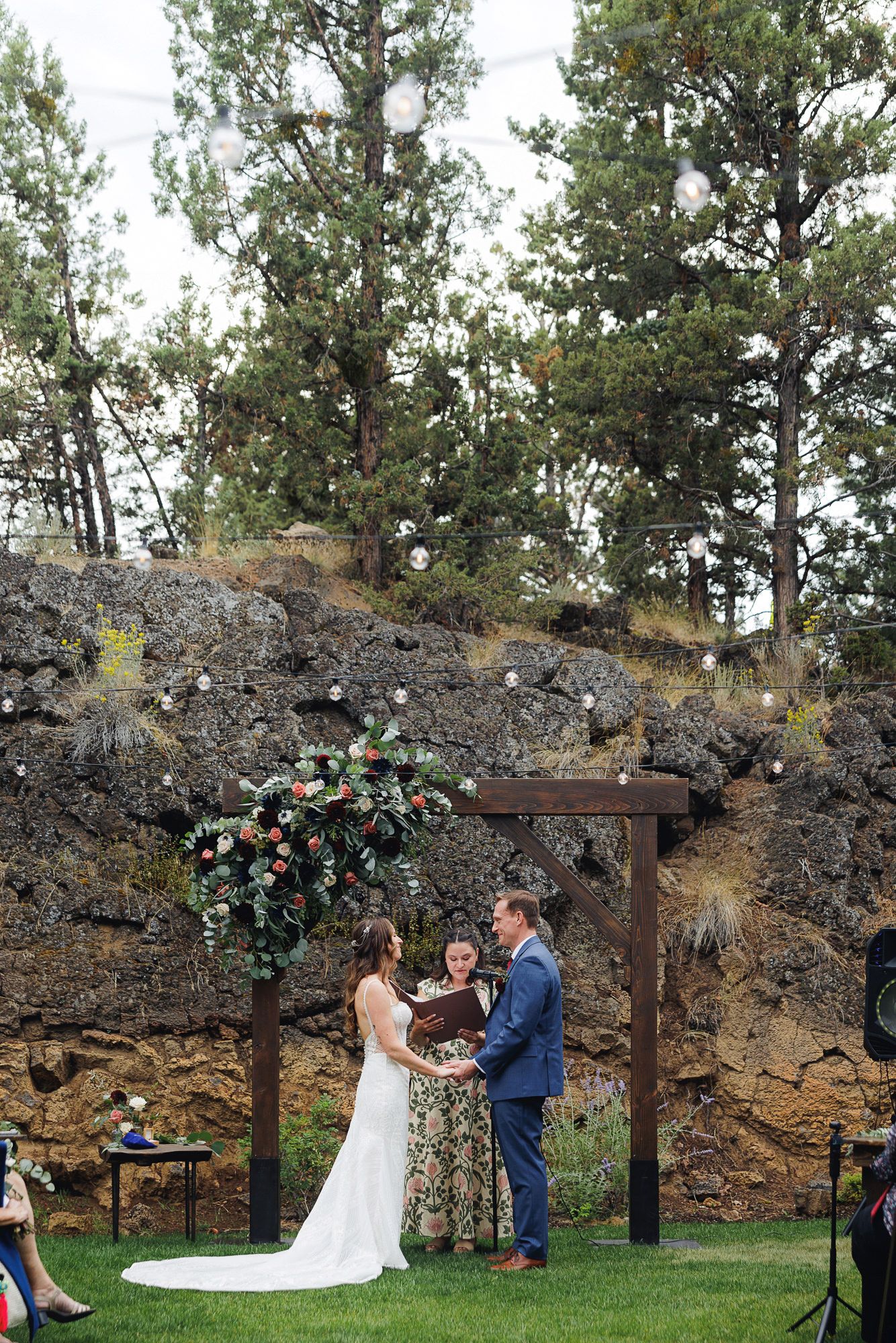 A couple exchanges vows during an outdoor wedding ceremony against a rustic stone wall backdrop with floral decorations.