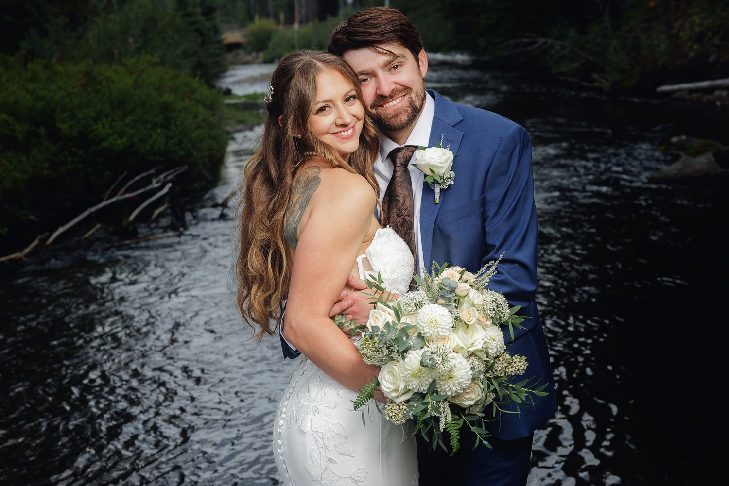 A couple in wedding attire embraces near a waterfall with white floral bouquet during their outdoor celebration.