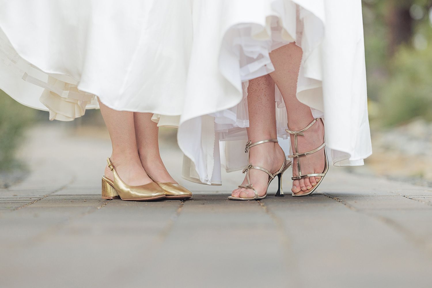 Close-up view of elegant bridal shoes and flowing white wedding dress hem on a stone path.