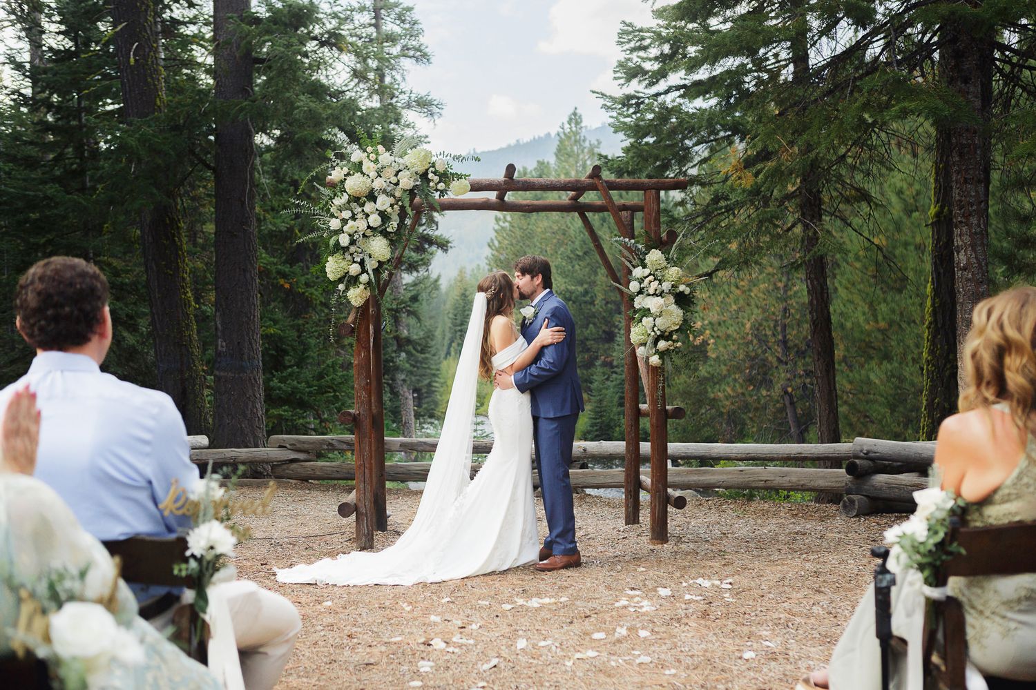 An outdoor mountain wedding ceremony takes place under a wooden arch decorated with white flowers in a forest setting.