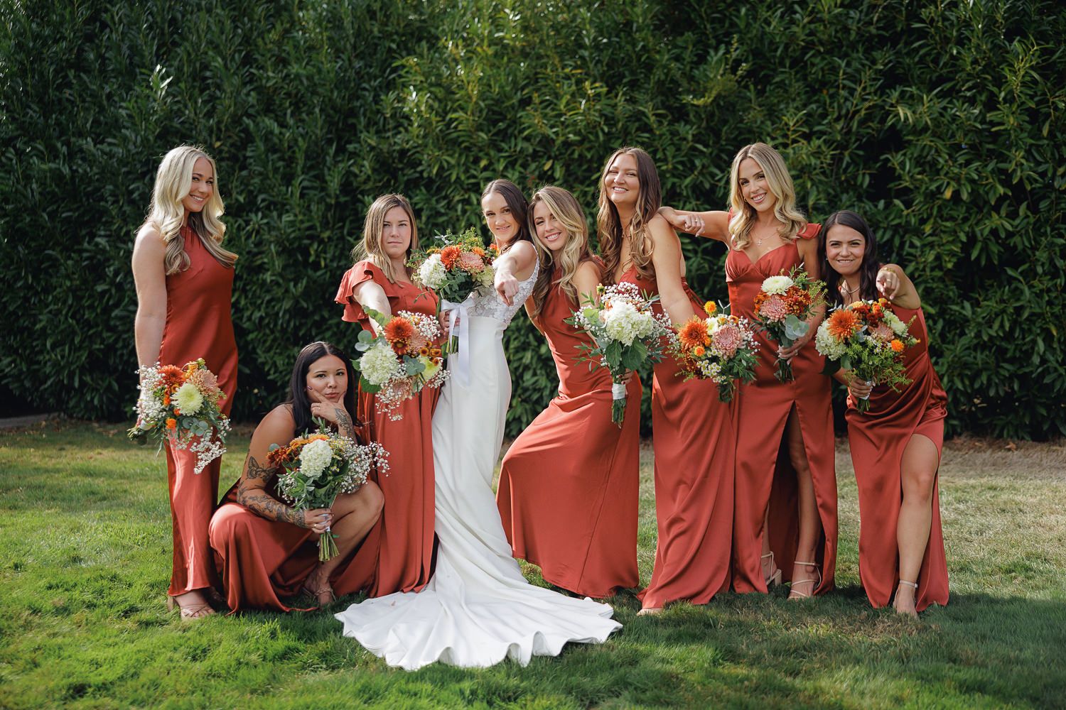 Wedding party in coral bridesmaid dresses poses together outdoors on grassy lawn for group photos.