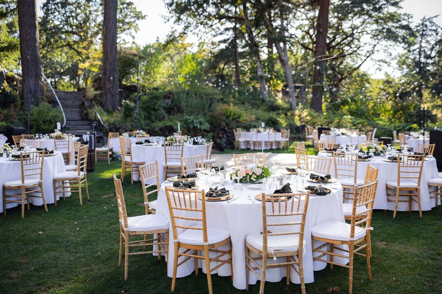Elegant outdoor wedding reception setup with round white tables and gold chairs arranged on a green lawn under tall trees.