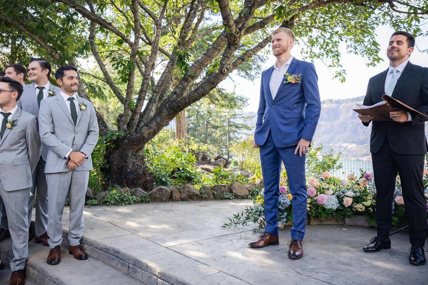 Wedding ceremony scene with groom and groomsmen standing under a large tree at an outdoor venue.