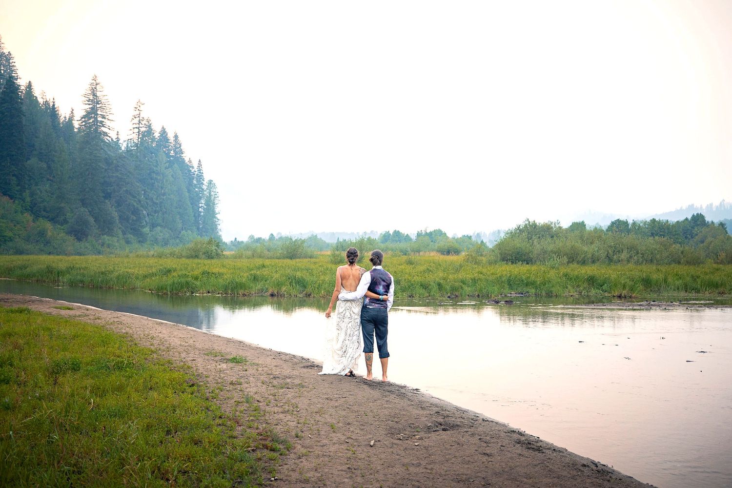 A couple stands together on a peaceful lakeside path surrounded by evergreen forest.