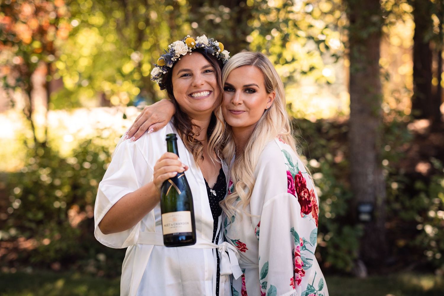 Two women in white robes smile together outdoors, one holding a champagne bottle, with sunlit trees in background.