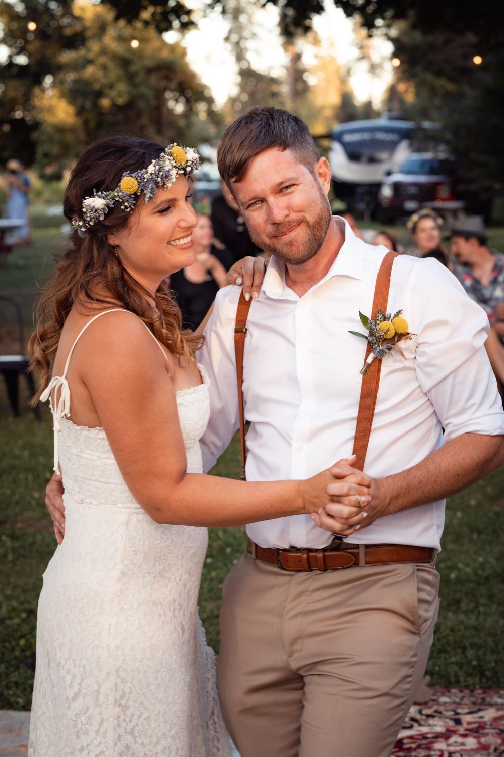 A couple dances during their rustic outdoor wedding celebration at sunset, wearing boho-style attire with floral accents.