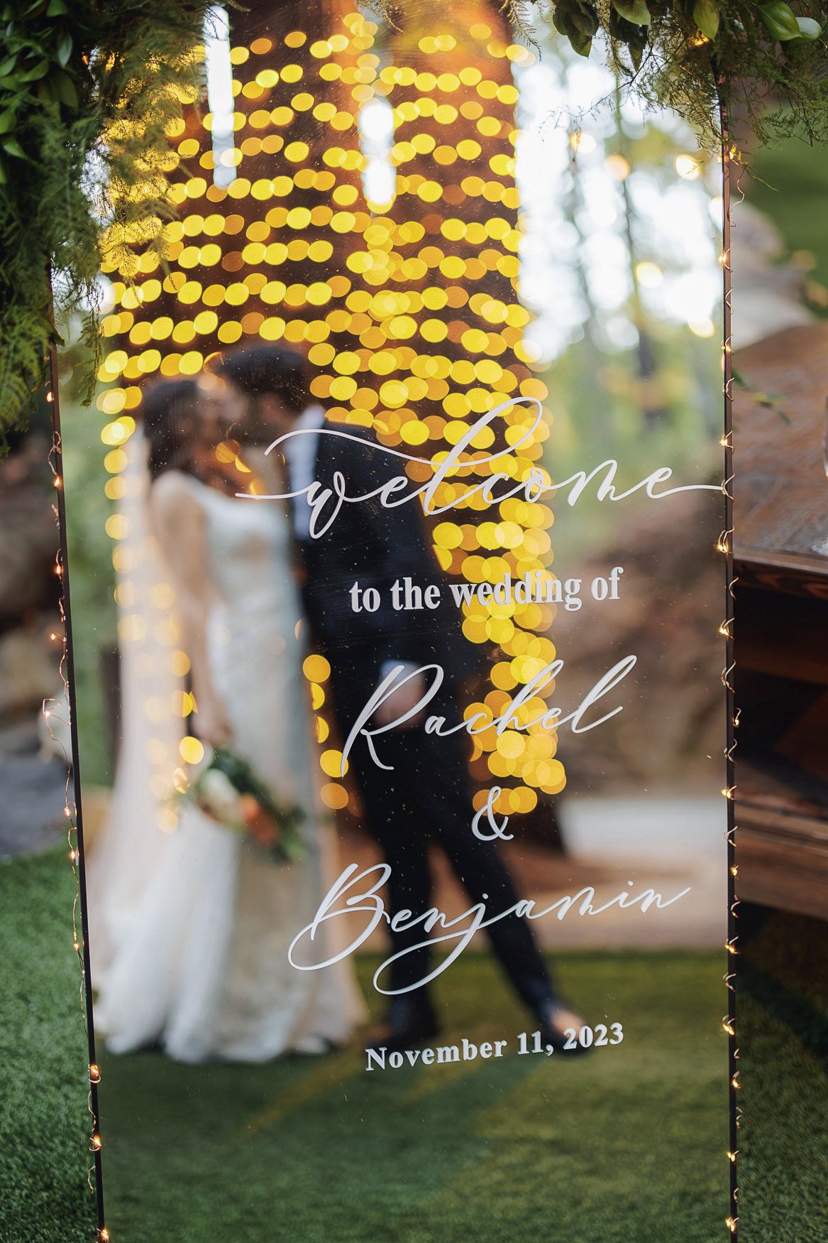 Elegant acrylic wedding welcome sign with blurred golden bokeh lights and greenery backdrop.