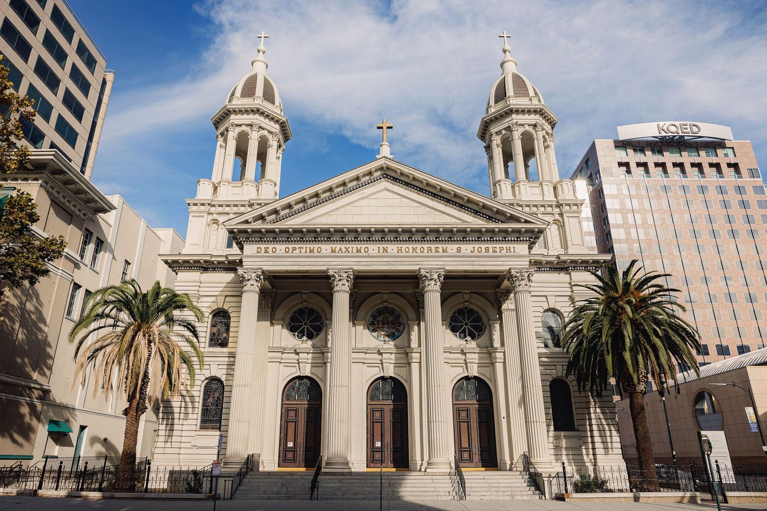 Historic San Jose church exterior with twin bell towers and neoclassical columns surrounded by palm trees and city buildings.