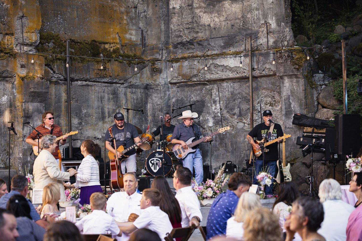 A rock band performs on stage in front of a concrete wall to a seated outdoor audience at a summer concert.