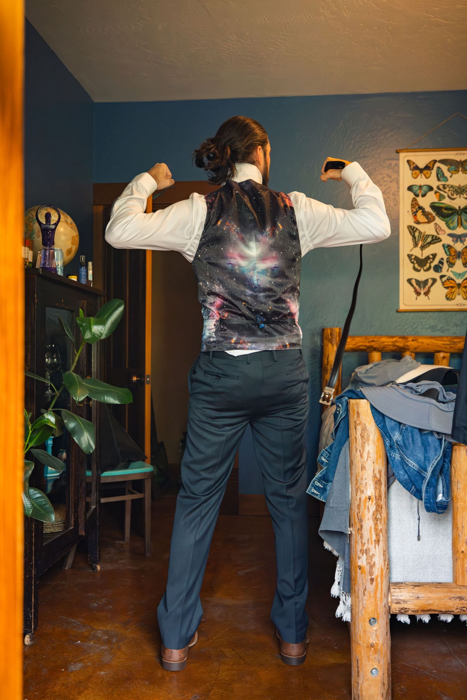 Person in formal attire showing off the back of a galaxy-patterned vest in a bedroom with houseplants and wall art.