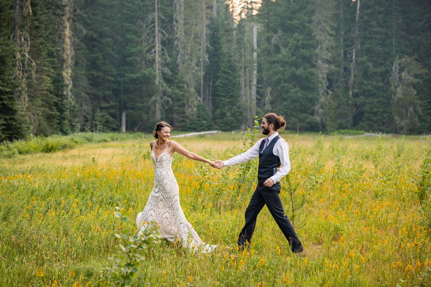 A couple in wedding attire holding hands while walking through a wildflower meadow surrounded by evergreen forest.