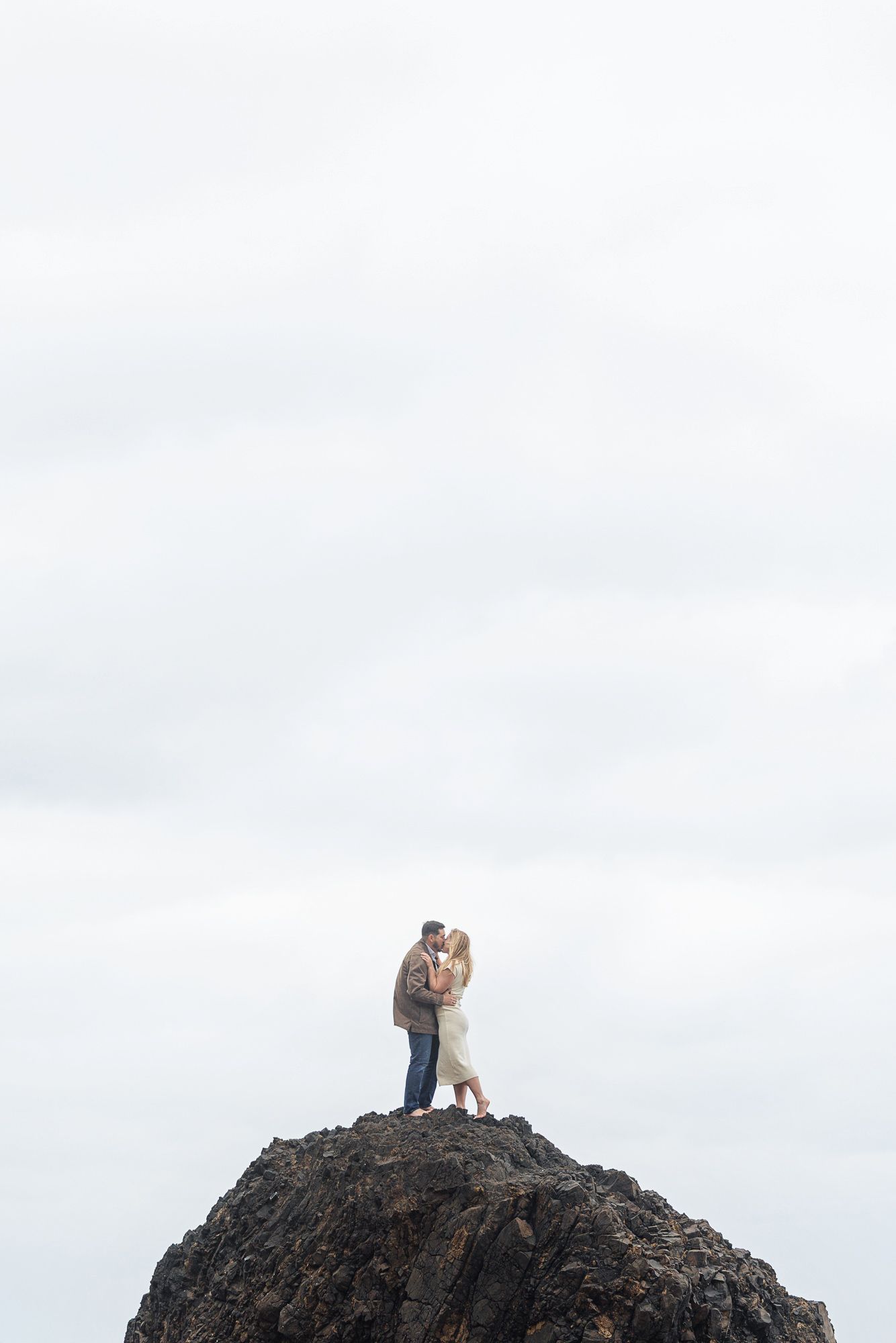 Couple kissing on top of a rocky cliff against a cloudy sky.