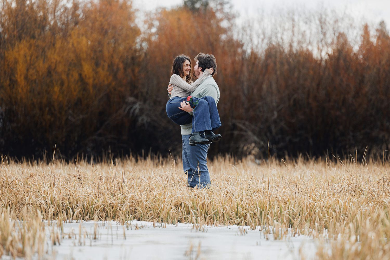 A couple embraces in a golden autumn field during sunset with tall grass and trees in the background.