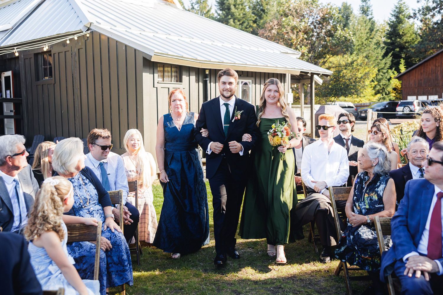 Groom walks down the aisle with bridesmaids in green dresses at an outdoor wedding ceremony.