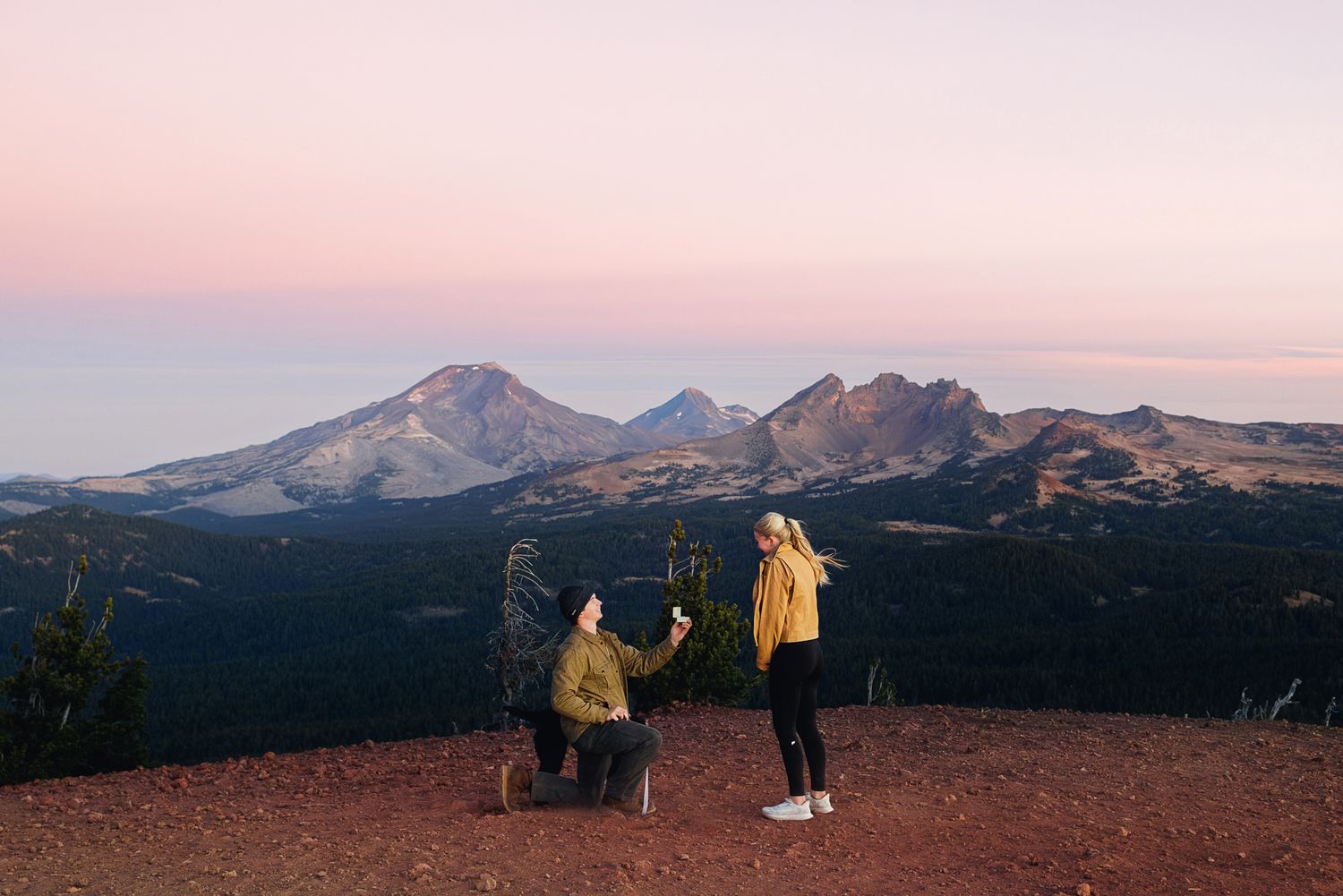 Person kneeling on one knee proposing to a standing partner on a mountain summit at sunset, with a snowy volcanic peak range behind them.