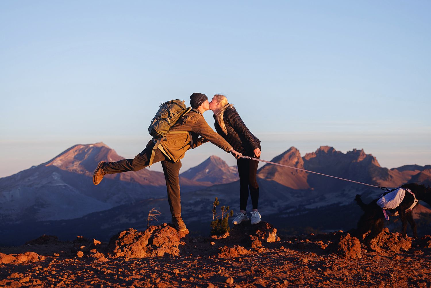 Hiker with a backpack leans in for a kiss with a partner on a rocky mountain ridge at golden hour, with rugged peaks in the background.