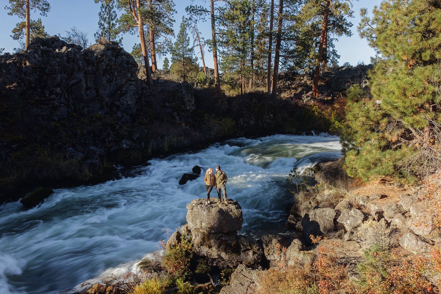 Two hikers stand on a large boulder beside rushing whitewater rapids, surrounded by dark volcanic rock cliffs and tall pine trees.