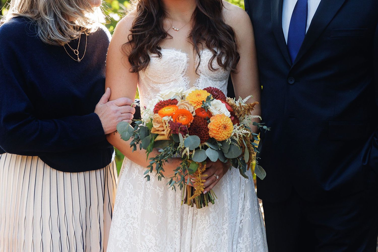 Bride in white lace gown holding autumn bouquet of orange and red roses with eucalyptus, flanked by wedding guests.