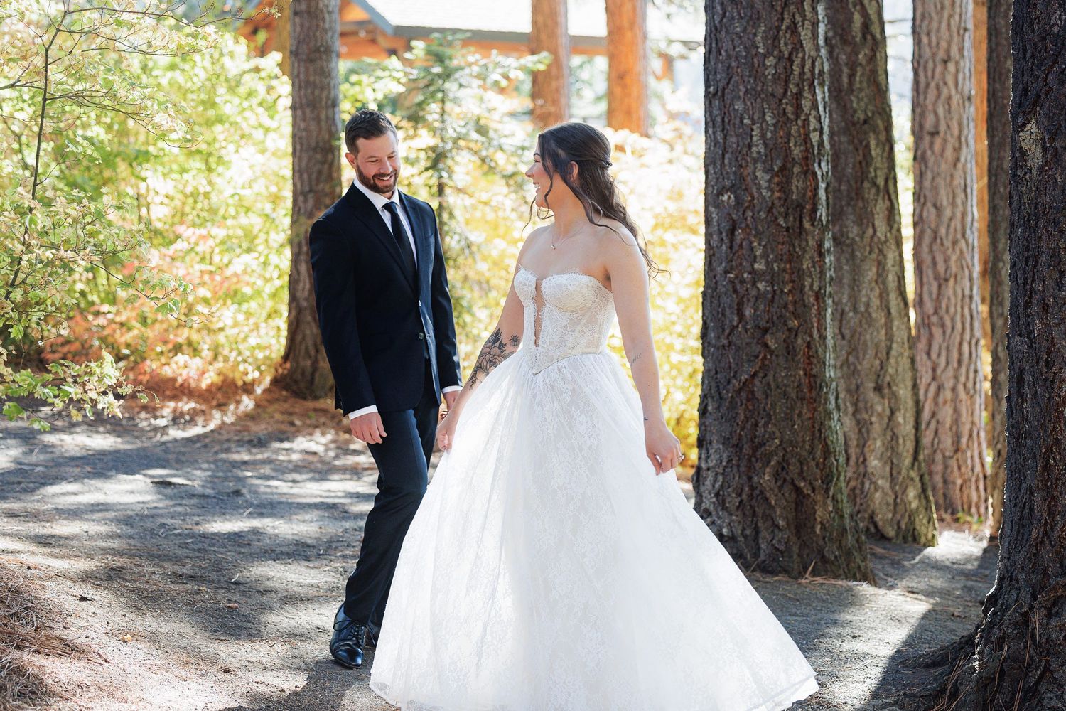 Bride in white ballgown and groom in navy suit share a romantic moment on a forest path surrounded by tall trees.