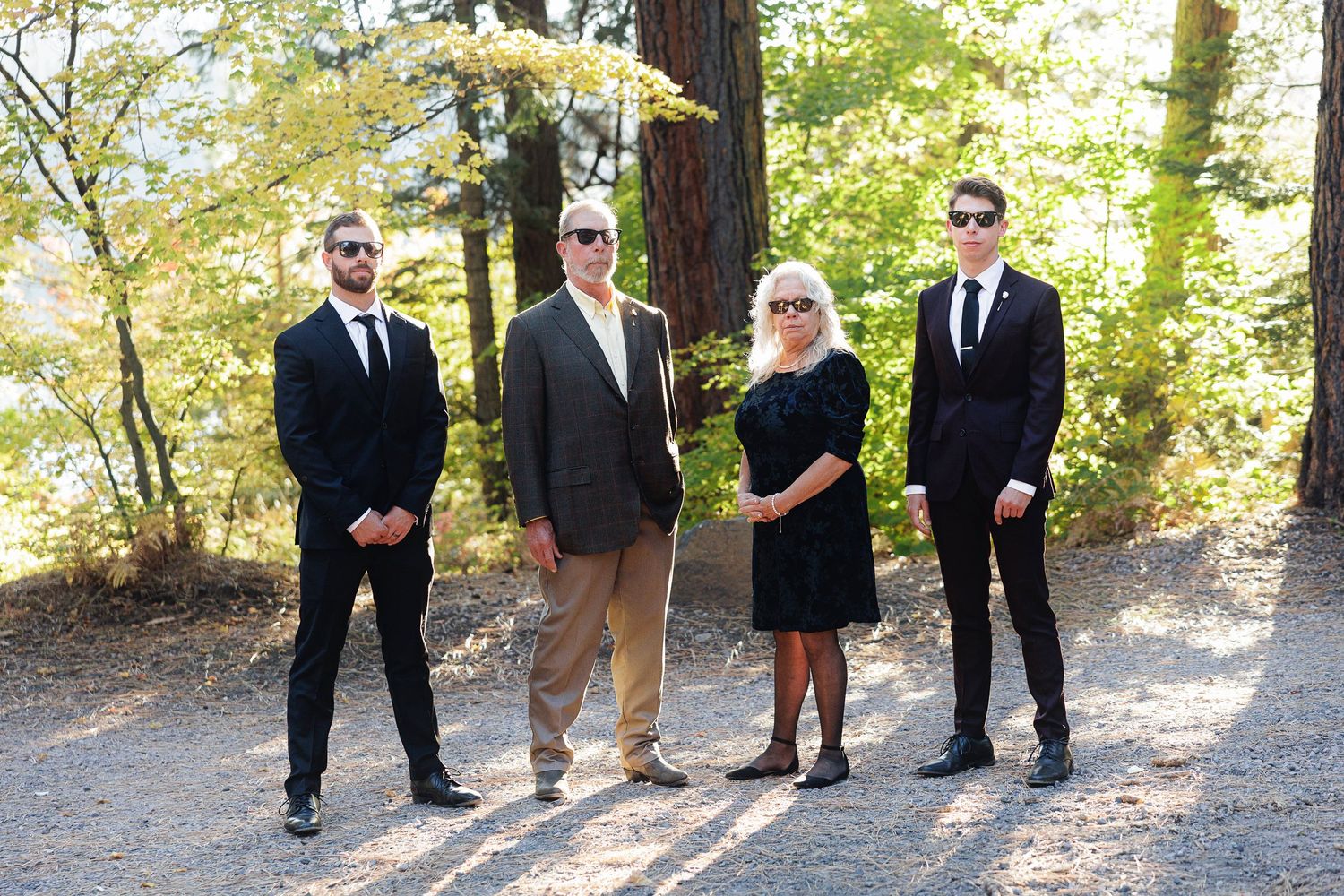 Family of four posing outdoors in formal attire on a gravel path surrounded by lush green trees.