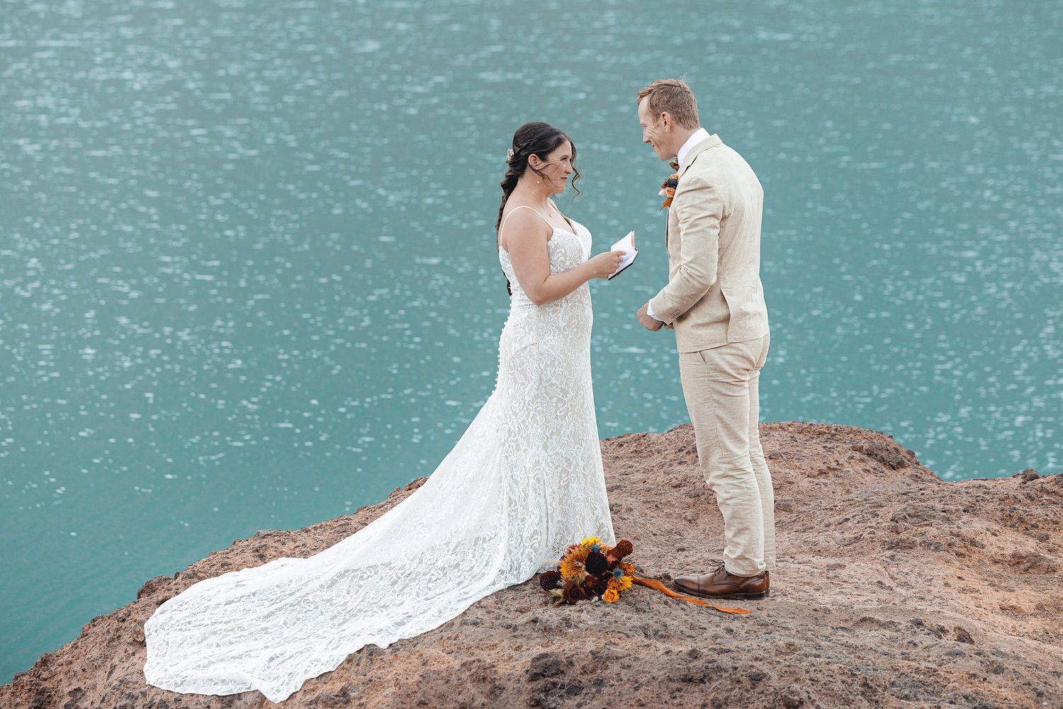 Bride in a lace wedding gown and groom in a beige suit exchange vows on a rocky cliff above turquoise water.