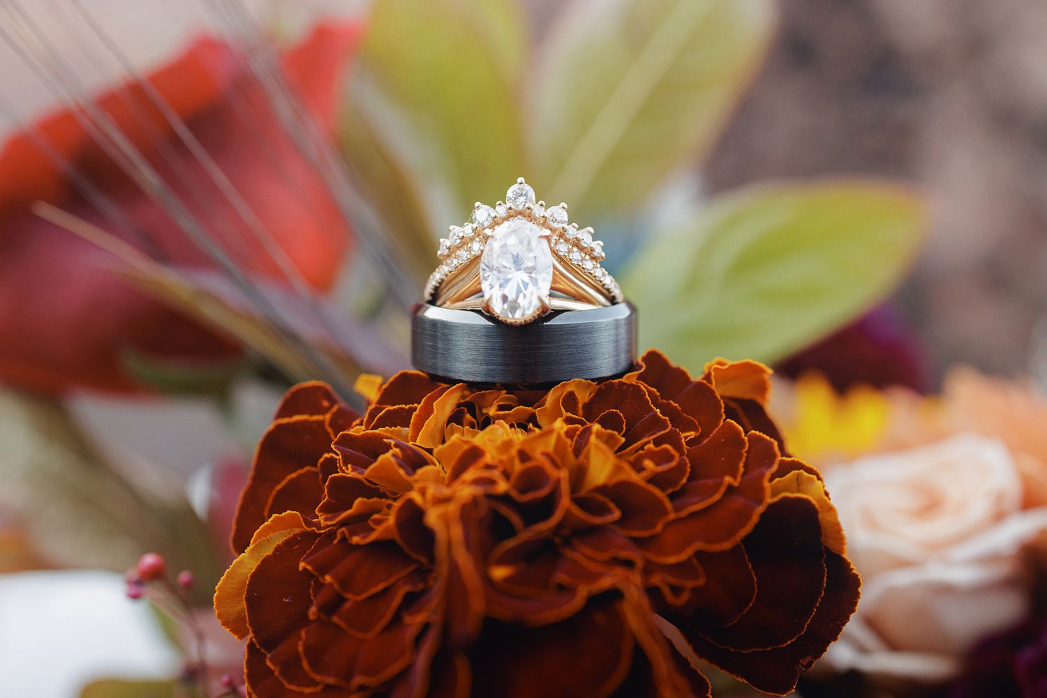 A diamond engagement ring with a gold chevron band rests atop a dark men's wedding band on a rust-orange marigold bouquet.