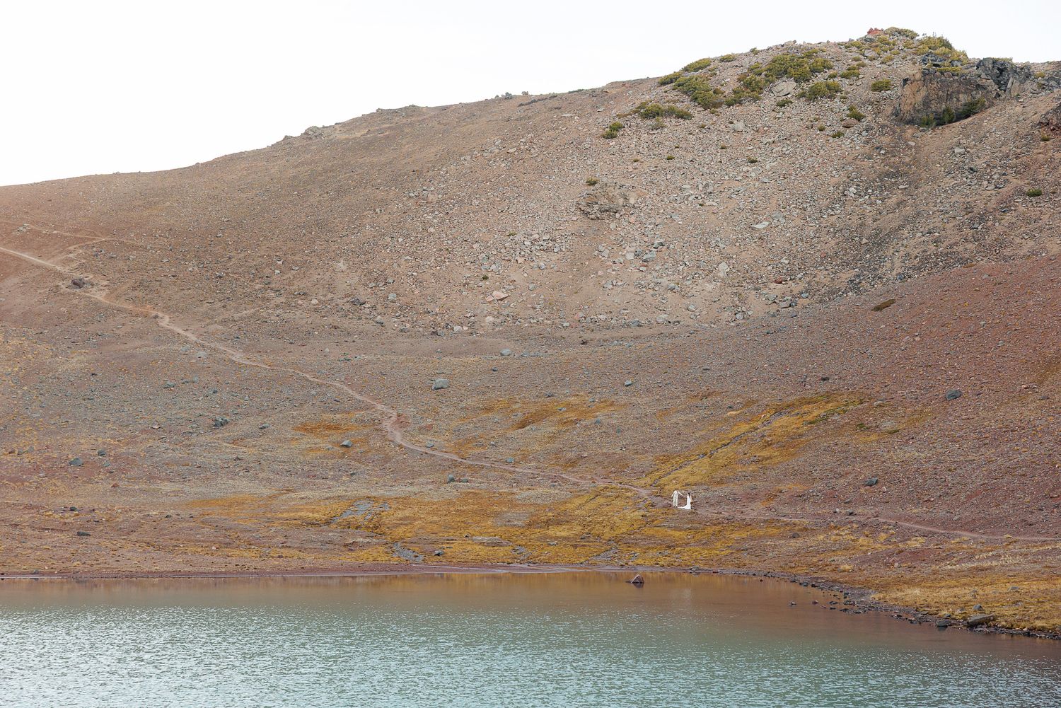 A small turquoise lake sits below a barren volcanic hillside with reddish-brown slopes, golden moss patches, and a faint hiking trail.