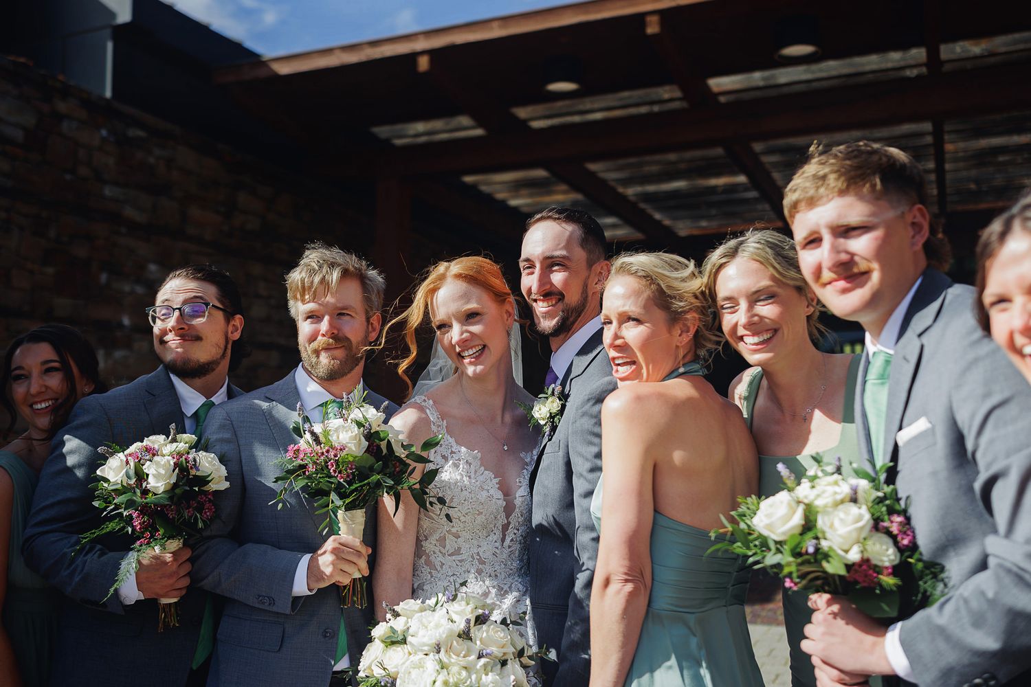 Wedding party smiling outdoors, bride in lace gown holding white bouquet, bridesmaids in teal dresses, groomsmen in gray suits.