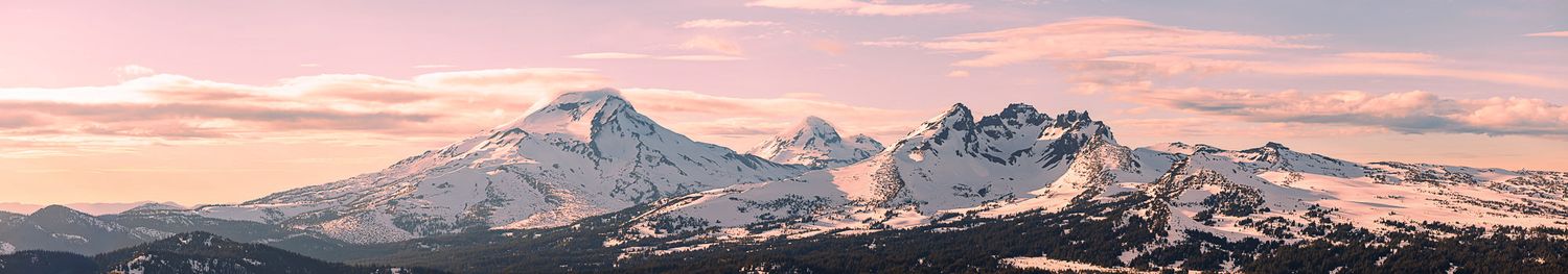 Panoramic view of snow-capped mountain peaks glowing pink and purple at sunset against a pastel winter sky.