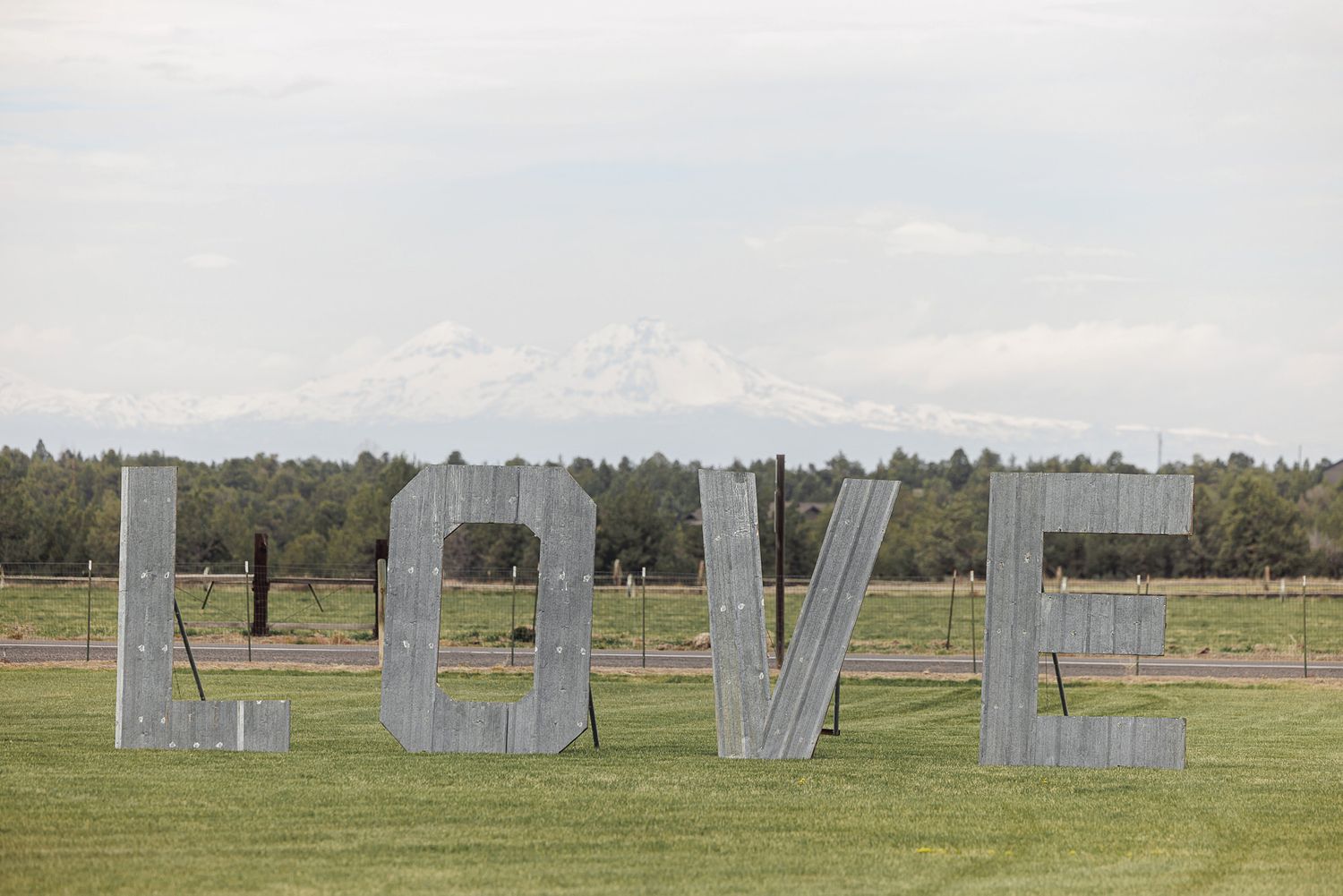 Large rustic metal LOVE letters on green grass with a snow-capped mountain and forest in the background.