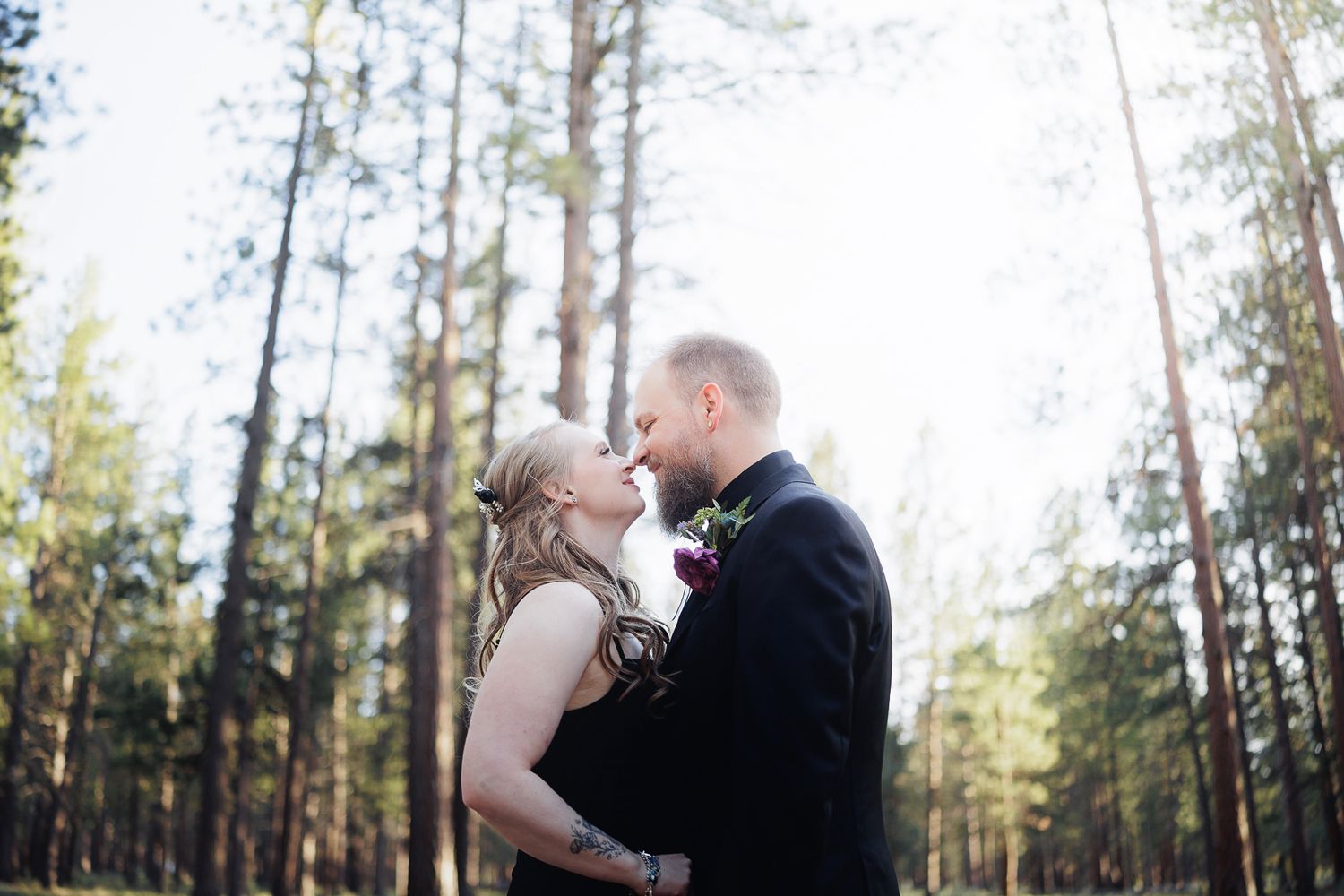 A bride and groom share a romantic moment in a sunlit forest, gazing into each other's eyes on their wedding day.