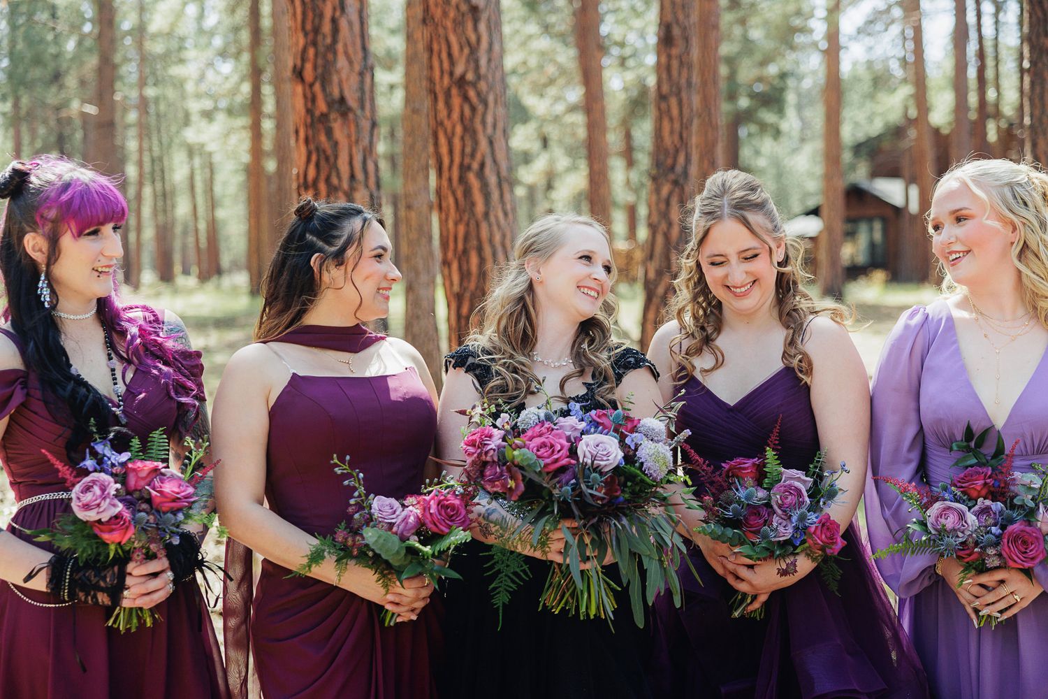 Bridesmaids in purple and plum gowns holding floral bouquets laughing together outdoors among tall pine trees.