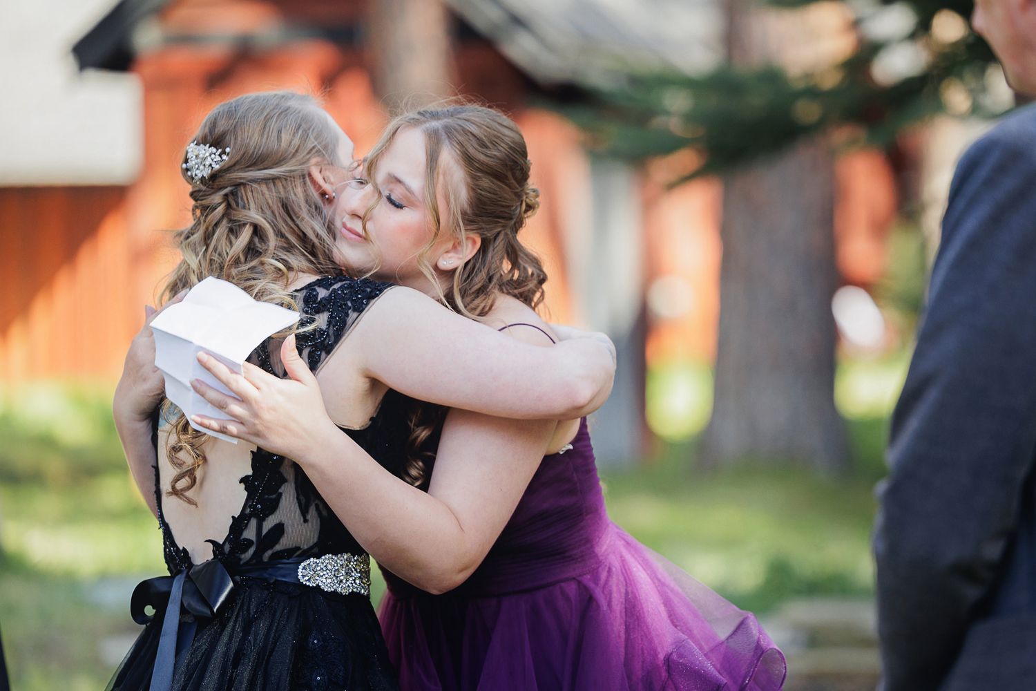 Two young women in formal dresses share a warm hug and kiss at an outdoor event.