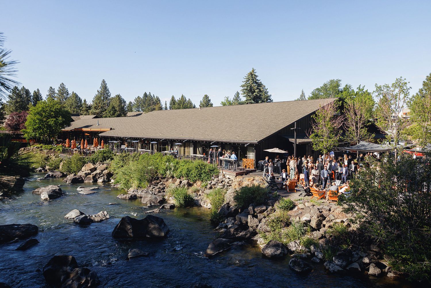 Rustic lodge beside a rocky river with guests gathered on the patio surrounded by tall pine trees at dusk.