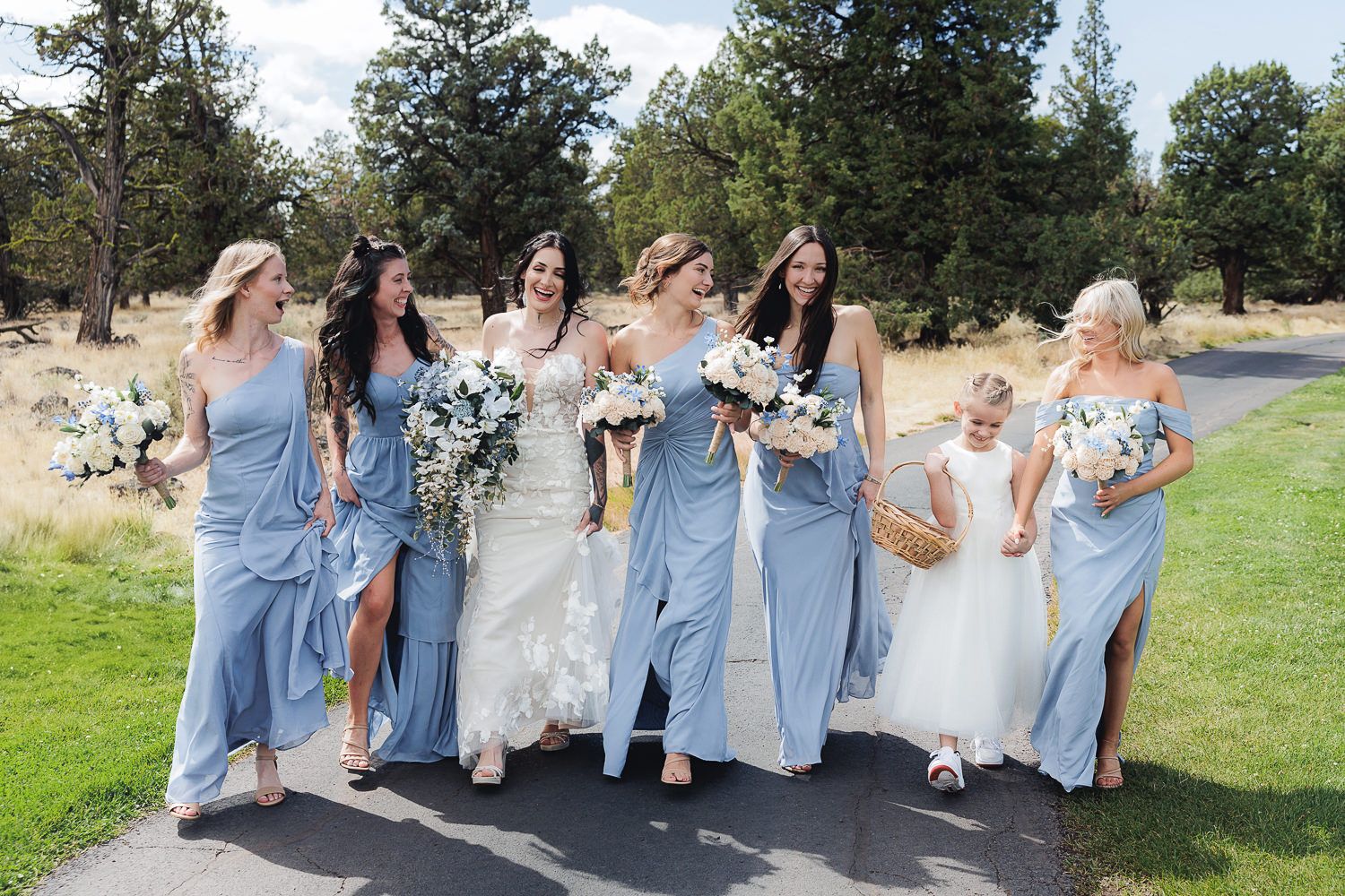 A bridal party wearing light blue bridesmaid dresses and white wedding gowns walk together on a path outdoors.